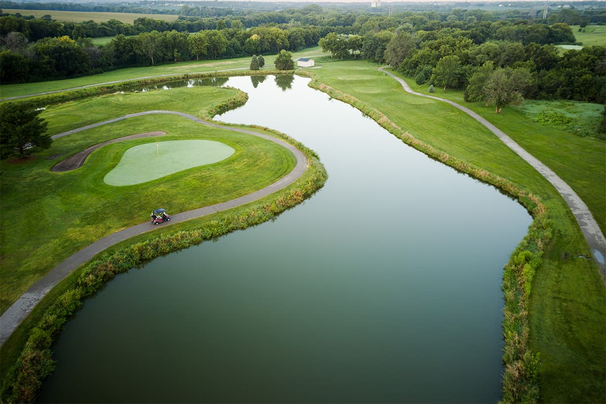 Aerial view of a Crooked Creek Golf Club hole featuring a large water hazard, manicured green, winding cart path, and scenic surroundings.